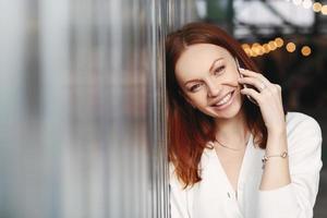 Photo of lovely woman with reddish hair, positive smile, has telephone talk with customer, calls to friend, dressed in white shirt, looks at camera with satisfied expression, enjoys communication