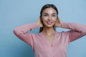 Portrait of adorable woman with perfect clean skin, toothy gentle smile, keeps hands on hair, wears rosy jumper, makes photo in studio, poses against blue background. Cosmetology and beauty concept