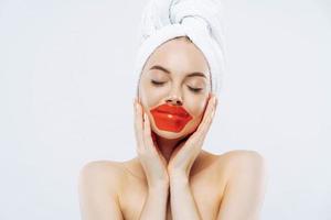 Cosmetology and skin care concept. Pretty woman with calm expression, closes eyes, applies lips patches, wears natural makeup, wrapped towel on head, stands bare shoulders indoor, white background photo