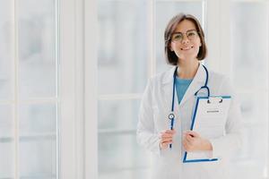 Beautiful smiling intern holds clipboard with written papers and pen, wears white uniform, ready to see patient in own private clinic, stands indoor against white background. Copy space area photo