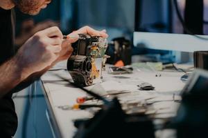 Woking process. Unknown man repairs professional digital camera, uses screwdriver, poses at workplace, holds photocamera disassembled part photo