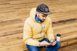 Hipster young bearded man wearing yellow jacket, stylish black cap and jeans sitting at wooden floor holding smartphone listening to music with earphones drinking takeaway coffee having relaxation photo