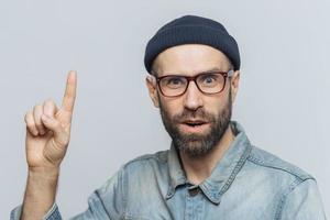 Amazed shocked man wears rectangular spectacles, looks with suprised expression, raises fore finger, has hush reaction, isolated over grey background. People, emotions and body language concept photo