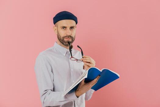 Self confident serious man with dark stubble, takes off spectacles, has attentive look at camera, holds opened textbook, learns necessary information, poses against pink background. Education photo