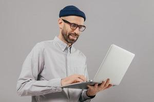 foto de un hombre satisfecho con barba gruesa y bigote, sostiene una computadora portátil, transfiere dinero, usa el servicio de banca en línea, envía archivos, se conecta a Internet inalámbrico, modelos sobre fondo gris