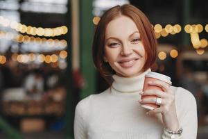 Photo of attractive young woman holds takeaway coffee, has pleased expression, toothy smile, dressed in white jumper, poses outside with blank space for your promotional content or advertisement