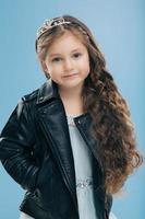 Photo of attractive woman with curly hair, has blue eyes, dressed in leather black jacket, keeps hands in pockets, models over blue background, wears crown on head. Children and fashion concept