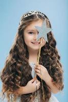 Isolated image of satisfied beautiful curly woman with blue eyes, covers face with magic wand, has long curly hair, poses over blue background, gentle smile on face, makes wish, dreams about something photo