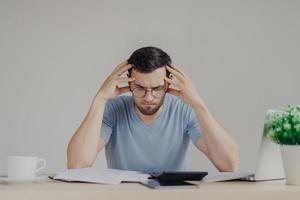 Brunet young male faces financial problems, not able to pay out mortagage, has desperate expression, makes calculations with calculator, isolated over grey background, uses modern technologies photo