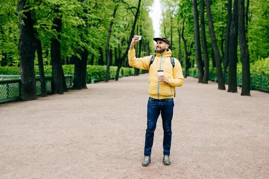 retrato horizontal de un apuesto hombre barbudo con anorak amarillo, gorra y jeans con expresión feliz mientras posa en el parque haciendo selfie caminando tomando café. hipster emocionado por selfie foto