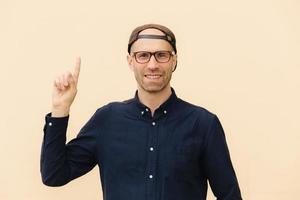 Cheerful male hipster with positive smile, keeps hand raised, indicates with fore finger upwards, dressed in fashionable clothes, wears spectacles isolated over light blank wall. Advertisement concept photo