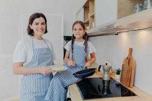 Horizontal shot of happy mother and small female child look gladfully at camera, busy preparing fried eggs for breakfast or dinner, pose together near cooker, being at kitchen. Family concept photo