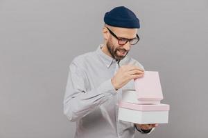 Glad young Caucasian man opens gift box, looks curiously, wears glasses, hat and white shirt, stands against grey background, celebrates something. People, special occasion and holiday concept photo