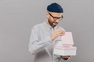 un joven caucásico alegre abre una caja de regalo, mira curiosamente, usa anteojos, sombrero y camisa blanca, se para en un fondo gris, celebra algo. personas, ocasiones especiales y concepto de vacaciones. foto