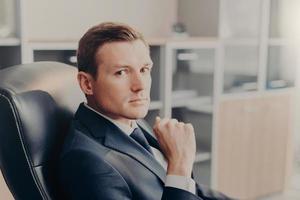 Professional male manager in formal suit, looks directly at camera, rests in chair at work place, conemplates about new business project. Elegant man director works in his own company or firm photo