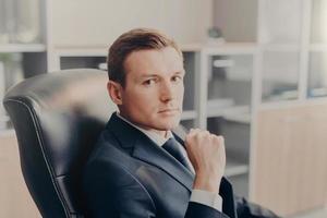 Indoor shot of serious confident businessman poses in chair, poses at cabinet, waits for colleagues to discuss main issues of work. Young male director in formal suit, thinks about new project photo