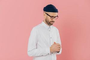 Contemplative unshaven man keeps gaze down, wears white shirt, hat and glasses, being deep in thoughts, tries to make solution, thinks how develop his business, isolated over pink background photo