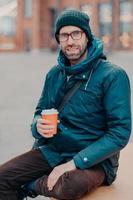 Stylish Caucasian man has free time, poses outside with disposable cup of coffee, dressed in hat and jacket, enjoys good rest, waits for someone at street, has satisfied expression. Lifestyle concept photo