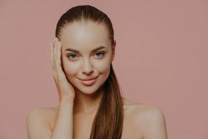 Isolated shot of pretty woman with combed pony tail, enjoys freshness of skin, looks with charming expression, stands bare shoulders, isolated on pink wall. Health care, makeup, spa and face lifting photo