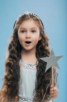 Small little child female princess wears crown and dress, holds magic wand, has long dark curly hair poses over blue background. Beautiful kid preapres for carnival or festive event. Childhood concept photo