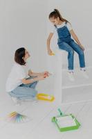 Shot of little girl sits on white furniture, refurbish drawer together with mother, use paint rollers and brush, pose in apartment against white background. Children, parents, repairing concept photo