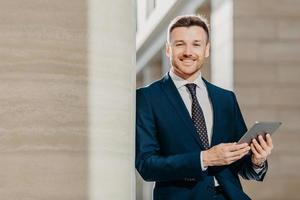 Horizontal shot of happy male manager dressed in black formal suit, uses modern tablet computer for making financial report, focused into distance with happy expression. People and business concept photo