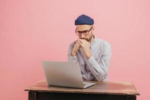 la toma horizontal de un trabajador masculino concentrado lee estadísticas atentamente, se enfoca en una computadora portátil, aprende algo en línea, usa anteojos, sombrero y camisa blanca, aislado sobre una pared rosa. foto