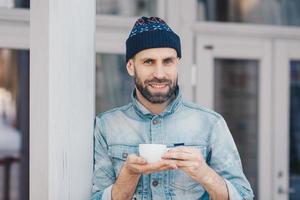 Portrait of attractive bearded male has pleasant smile, dressed in denim jacket, holds white cup of coffee or tea, enjoys good time, has coffee break. Happy handsome stylish young hipster guy photo