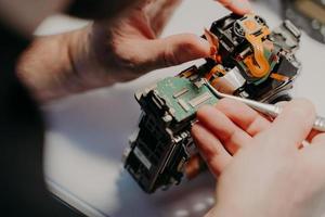 Hands of handyman repairing broken digital camera, holds special tweezers, works in workshop. Maintenance, occupation and repairing concept. photo