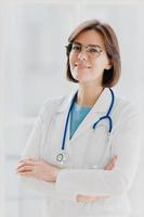 Shot of confident brunette female doctor or surgeon stands with hands crossed, wears white uniform, cares about health of patients ready to give advice any time. People, occupation, profession concept photo