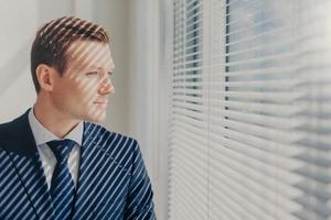 Prosperous male boss in elegant luxury suit, stands in modern office near big window with shutter, has rest after meeting with shareholders of company, thinks about something. People, work concept photo