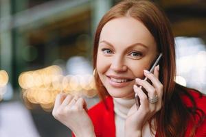 Close up shot of attractive carefree female student talks on mobile phone, enjoys conversation with groupmate, has pleasant smile, poses over blurred background, has spare time after classes photo