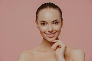 Studio shot of pretty charming woman touches chin, has healthy glowing skin, cares about complexion, stands naked indoor, has cosmetology procedure, isolated on pink background. Beauty concept photo