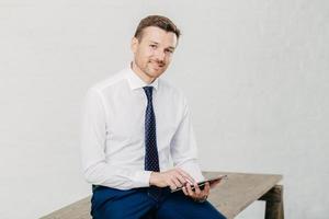 Happy male CEO makes online transaction on modern tablet computer, dressed in formal wear, poses on wooden table, has friendly expression, isolated over white background. People and work concept photo