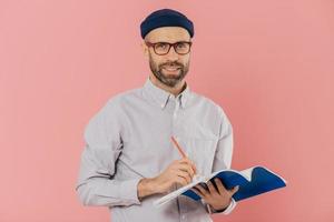 Pleased attractive man with stubble, wears transparent glasses, fashionable hat and shirt, writes with pen in notepad, prepares for speaking in front of auditorium, isolated over pink background photo