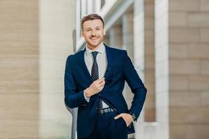 Waist up portrait of successful male lawyer comes on meeting with client, wears elegant formal clothes, holds shades, looks happily at camera, keeps hand in pocket. Business and people concept photo