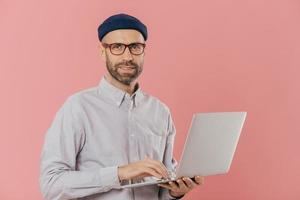 Isolated shot of handsome unshaven male does distance job on portable laptop computer, wears hat and optical glasses, connected to wireless internet, stands over pink background, develops software photo