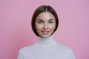 Isolated shot of charming glad young woman with pleasant smile, wears white turtleneck, looks directly at camera, isolated on pink background. Beautiful female student returns home from lectures photo