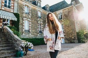 Outdoor portrait of pensive thoughtful beautiful woman with long staight luxurious hair keeps hands in pocket of mantlet, looks down, stands against ancient castle or builduing, wonderful exterior photo