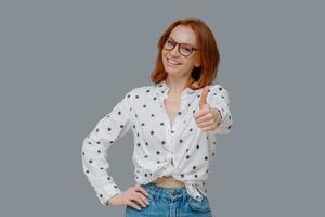 Pleasant looking confident woman with ginger hair, shows thumb up, says very good and encourages friend, likes someones idea, gives positive reply, wears stylish outfit, stands indoor over grey wall photo