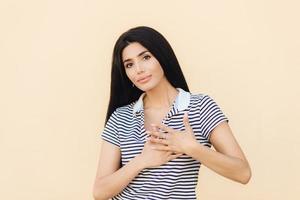 Friendly looking brunette female keeps hands on heart as expresses her gratitude, has serious expression, wears casual clothes, isolated over studio background. People, body language concept photo