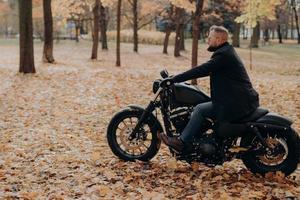 Horizontal shot of bearded biker in action or movement against park during autumn time, going to reach destination quickly, wears sunglasses, jacket, jeans and shoes, has adventurous trip alone photo