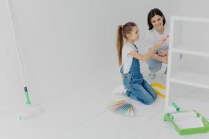 Photo of little girl and her mum paint furniture for new apartment, pose in empty room, use color samples, use painting tools, enjoy spending time together. House renovation and design concept