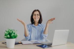 Happy overjoyed female achieves success in her work, going to present high results, raises hands joyfully, sits at working place with laptop computer, diary, calculator, isolated on grey background photo