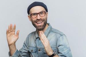 Portrait of happy middle aged man has fun indoor, raises hands, has pleased expression, wears glasses, hat and denim shirt, isolated over white background. People, positive emotions and feelings photo