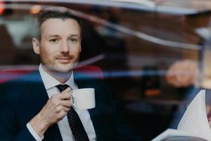 Attractive businessman in formal suit, drinks coffee during dinner break, sits in luxury cafe, looks thoughtfully at window, contemplates about business affairs has pleasant look. People and lifestyle photo