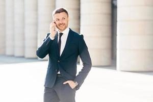 Portrait of successful male manager satisfied with mobile tariffs, makes phone call, uses banking service operator, wears formal suit, stands in urban setting outsie. Professional man worker photo