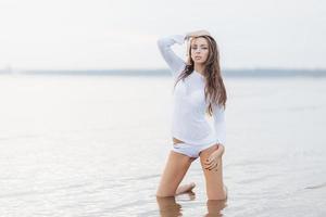 vista al aire libre de una guapa morena con maquillaje, vestida con bikini blanco, posa frente a la cámara cerca del fondo del mar en calma, tiene forma de cuerpo en forma, se siente libre y relajada. mujer en la orilla del mar foto