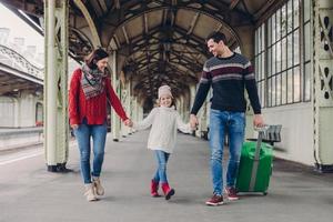 Three family members on railway station. Happy mother, daughter and father have positive facial expressions, waits for train on platform, enjoy togetherness. Glad passengers have journey abroad photo