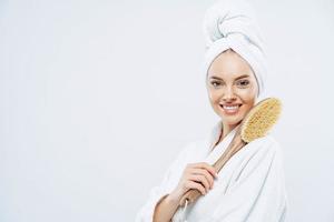 Sideways shot of beautiful young European woman with charming smile, poses with bath item, uses brush to massage body, poses against white background, blank space for your promotional content photo